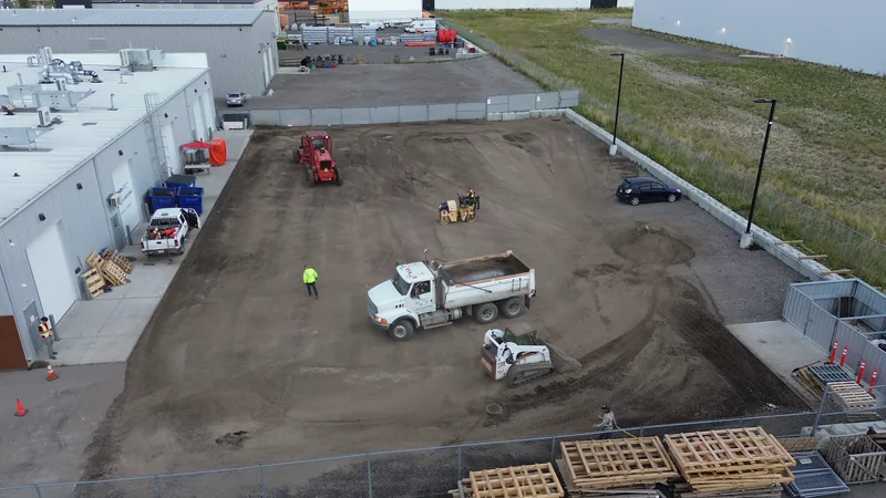 Aerial of a fenced compound lot during base prep: dump truck, skid steer and roller on the graded gravel