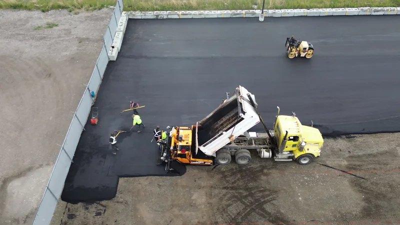 Dump truck feeding the paver as the first hot mix asphalt mat goes down along a compound wall