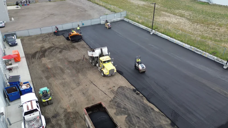 Aerial of a fenced compound lot in SE Calgary half paved, with the dump truck, paver and roller at work
